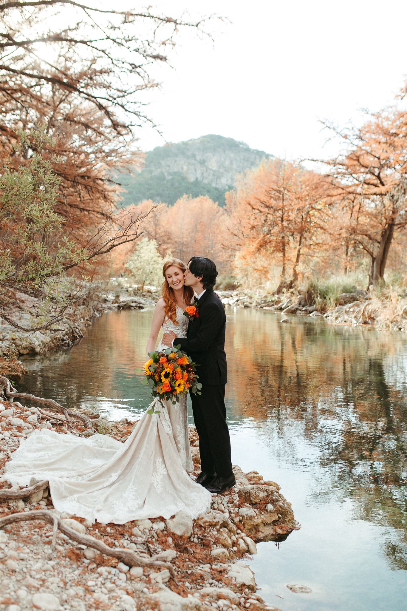 bride and groom posing next to river with fall trees hill country wedding photographer leah thomason