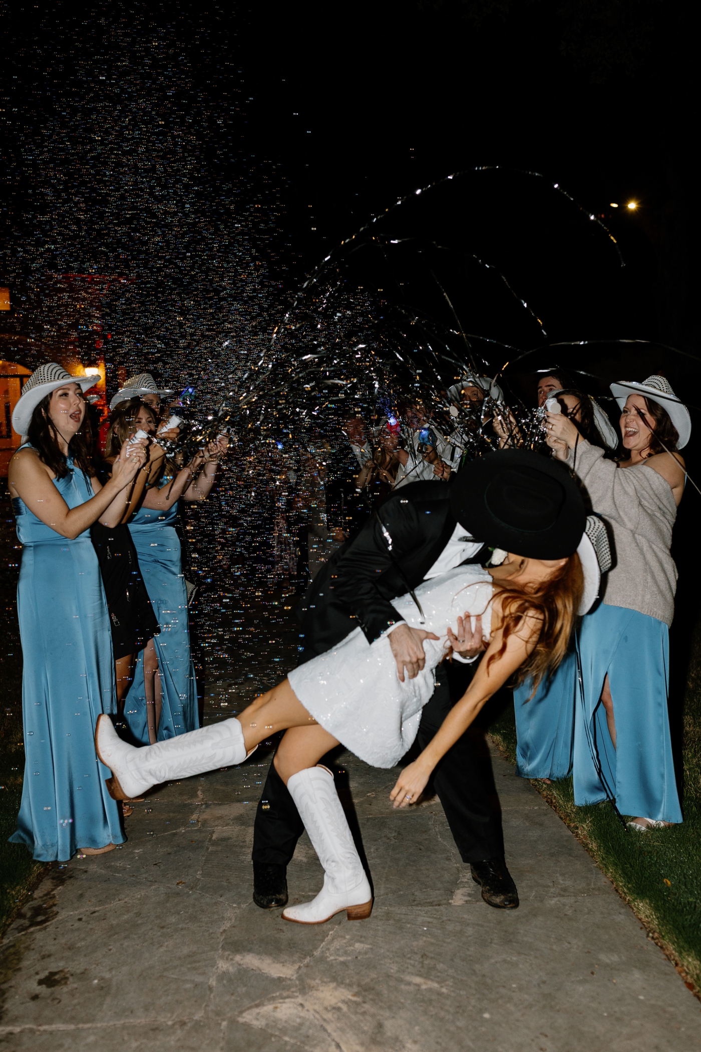 A bride and groom kiss while thier wedding guests blow bubbles and wave streamers