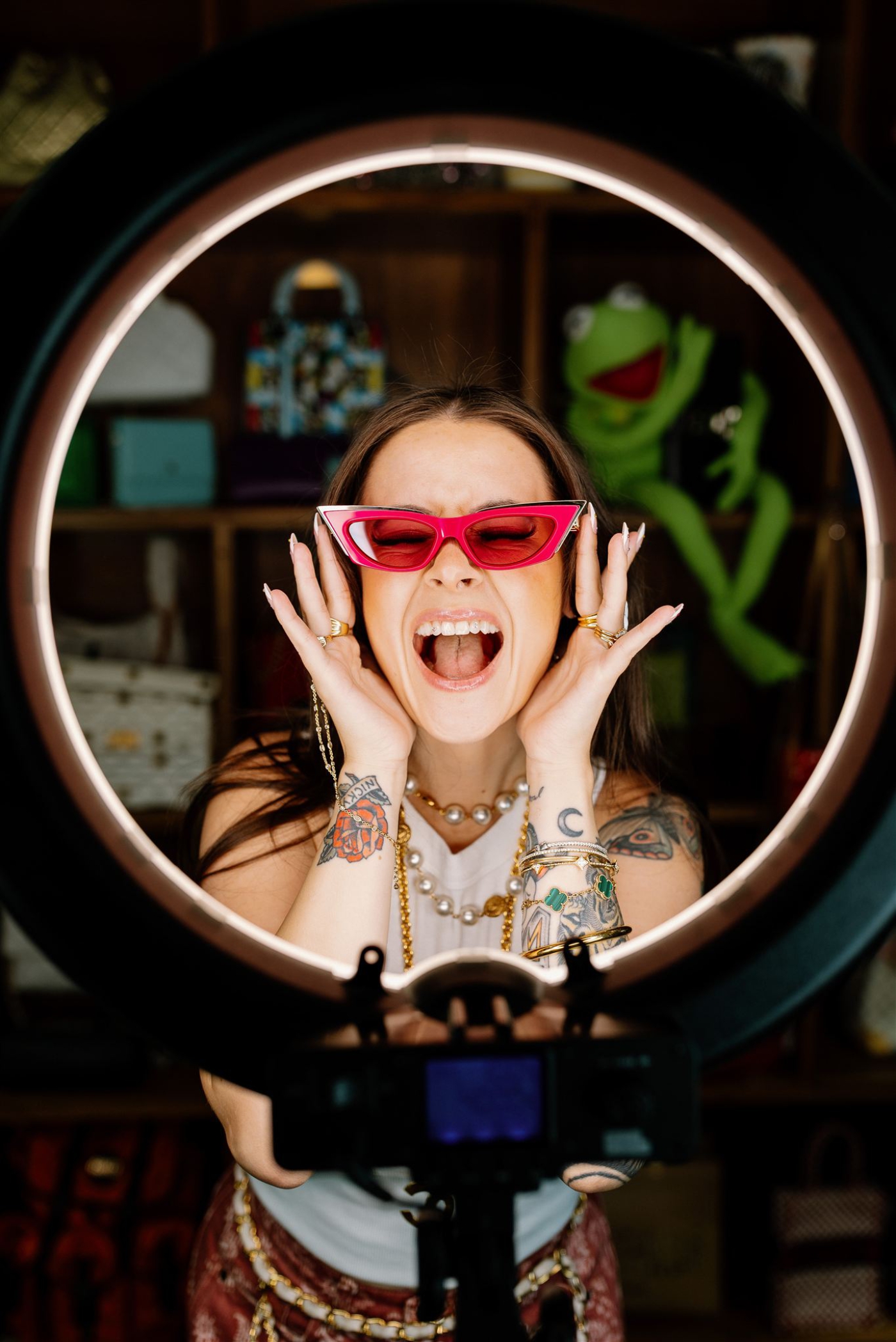 A woman stands in front of a ring light with retro red sunglasses on