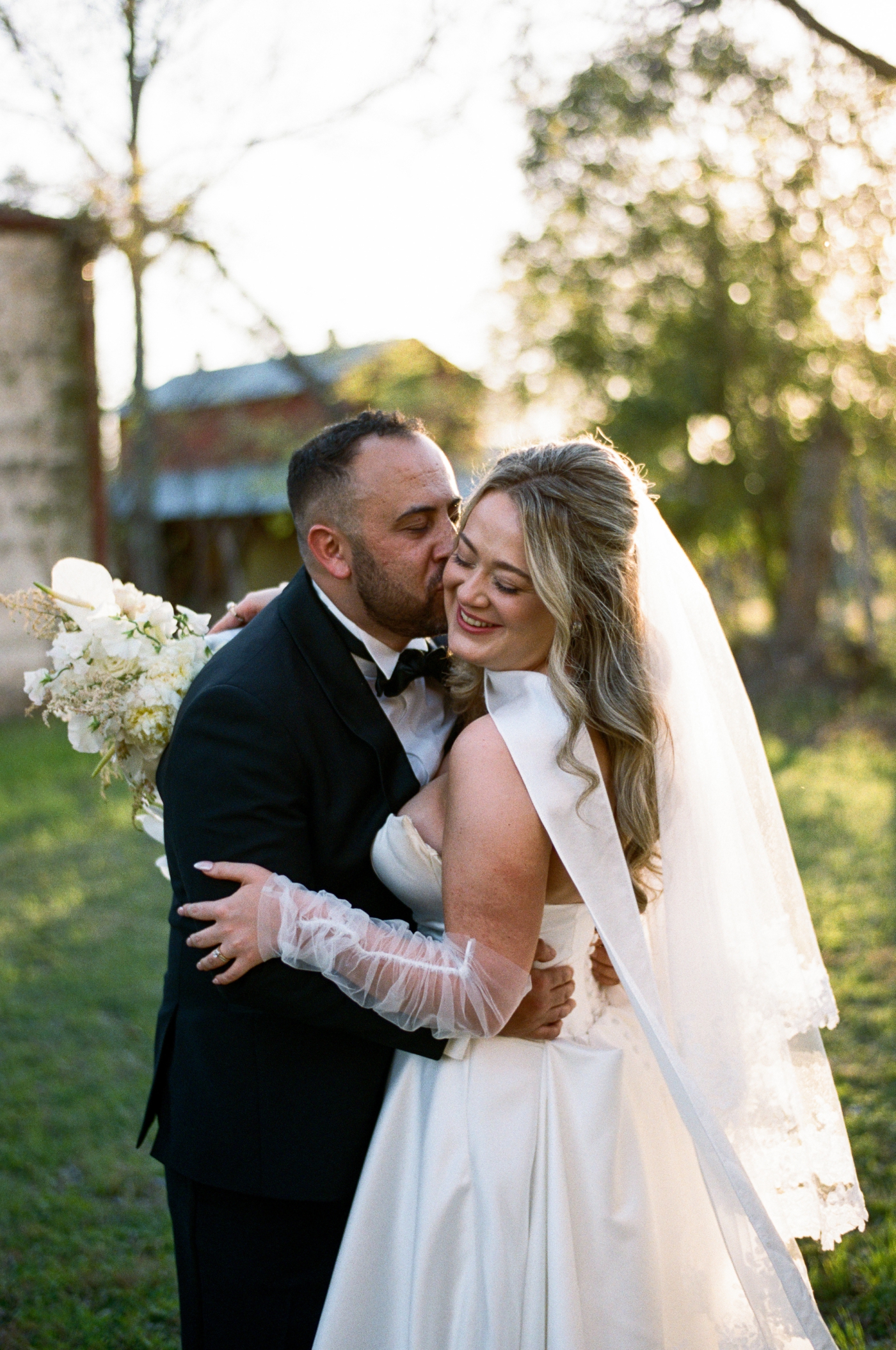 A bride and groom embrace while golden light shines on the bride's veil